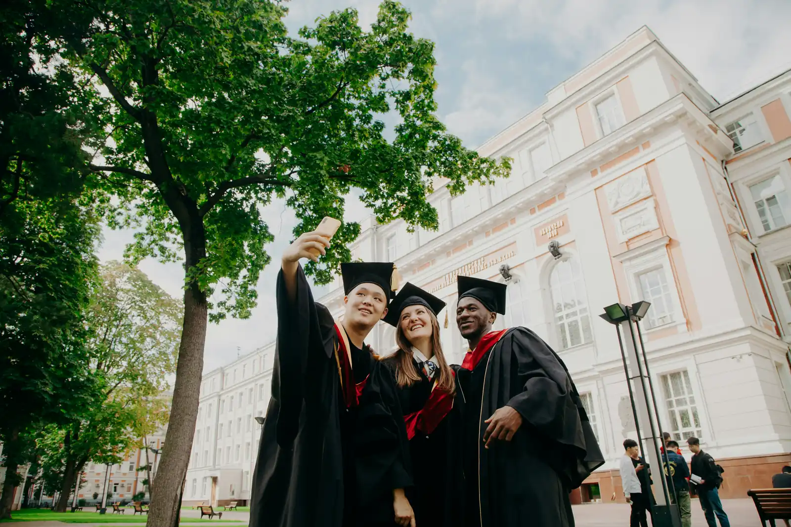 University graduates in caps and gowns taking a selfie outside a grand campus building